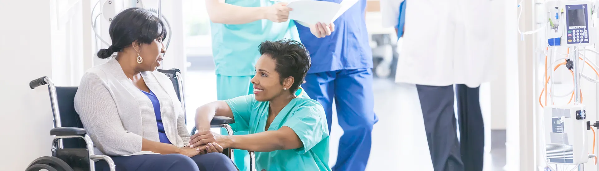 A black woman in a wheelchair smiling while a black nurse holders her hand while kneeling down to talk to her.