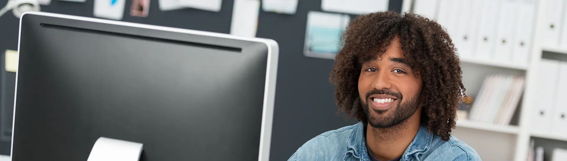 A black man with curly long hair smiling while sitting behind a computer monitor.