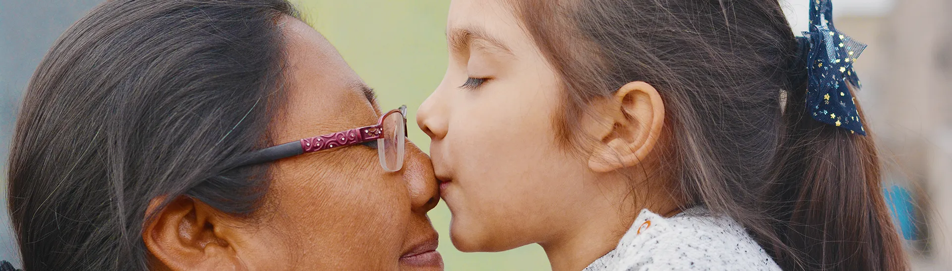 A young girl is giving a kiss to an elder woman (maybe her grandmother) on the nose.