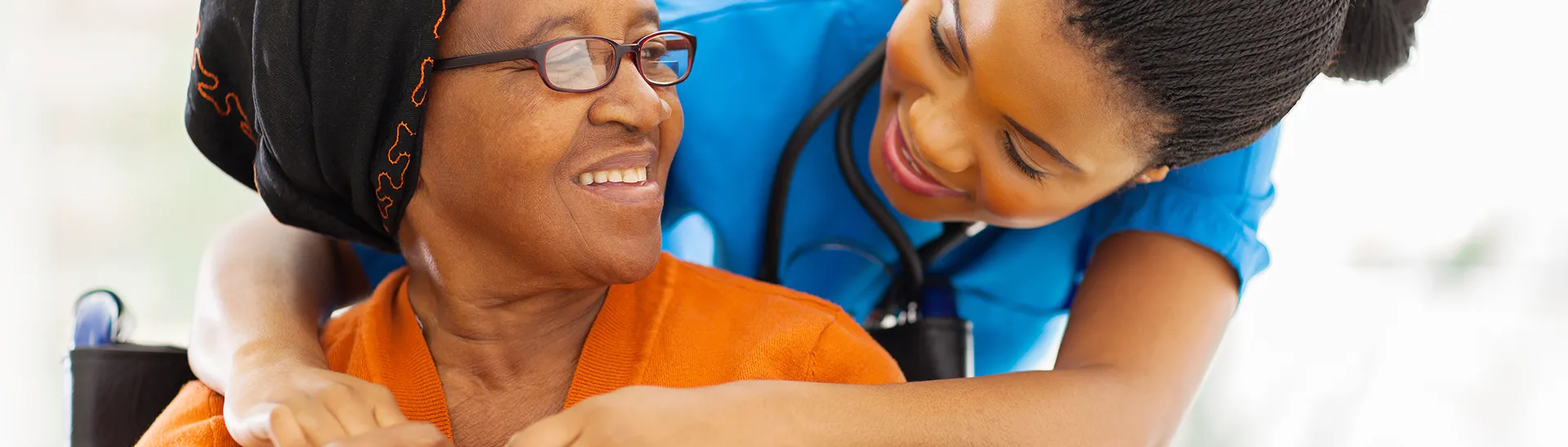 A younger nurse with her arms wrapped supportively around an elder woman's shoulders while she sits.