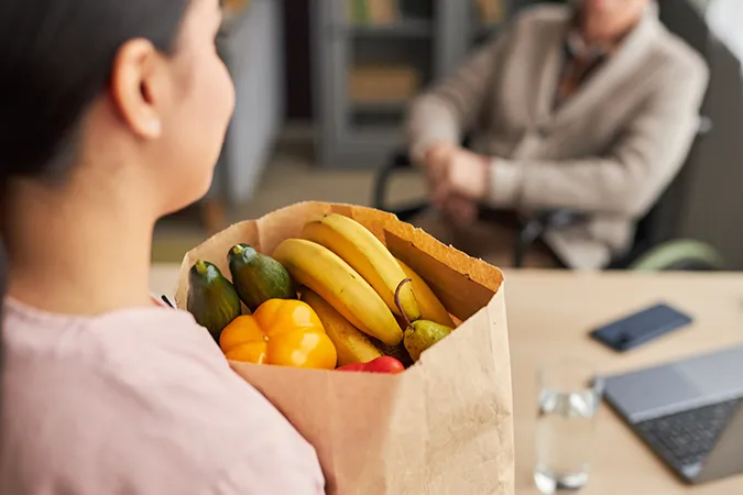 Woman-Holding-Groceries_2261597103 A woman holding a paper bag full of fruits and vegetables while talking to a woman in a wheelchair.